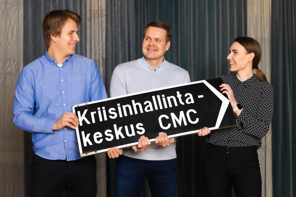 Two men and one woman are standing and supporting, smiling, facing a sign that reads the Crisis Management Center CMC. In the background a blue-gray curtain.