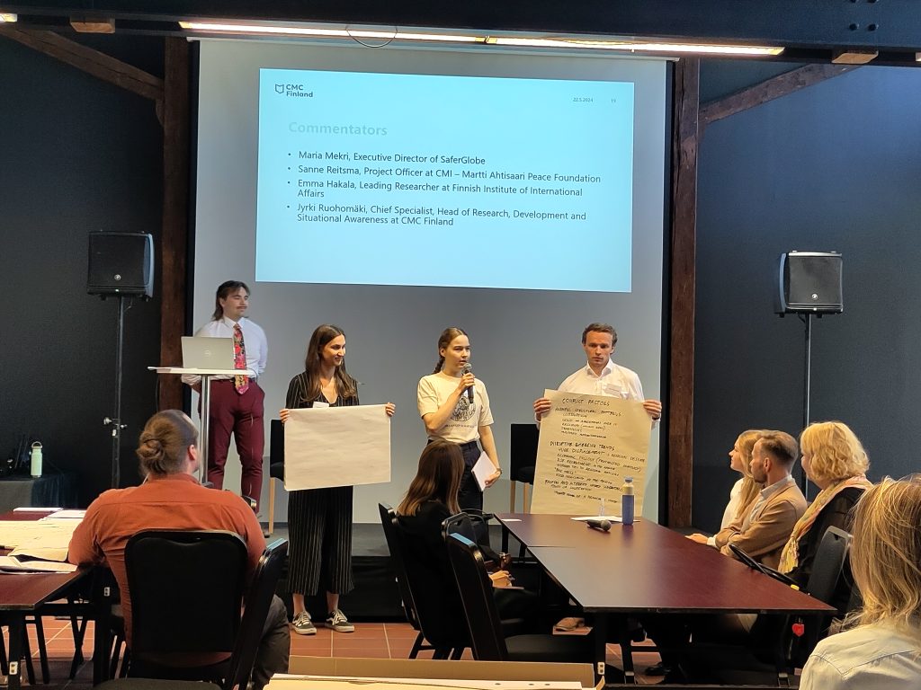 Three people are standing. One of them speaks on a microphone and two of them are holding large papers which is the group's conflict analysis. The audience is listening to their presentation.